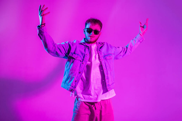 Créatif beau danseur de mode homme avec des lunettes de soleil en denim élégant danse dans un studio coloré avec lumière rose et néon - Photo, image