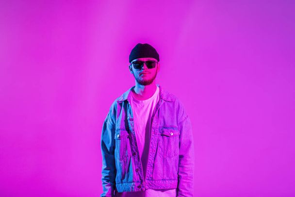 Stílusos jóképű, kreatív, fiatal srác sapkával és napszemüveggel divatos farmerdzsekiben egy rózsaszín és neon fényű stúdióban - Fotó, kép