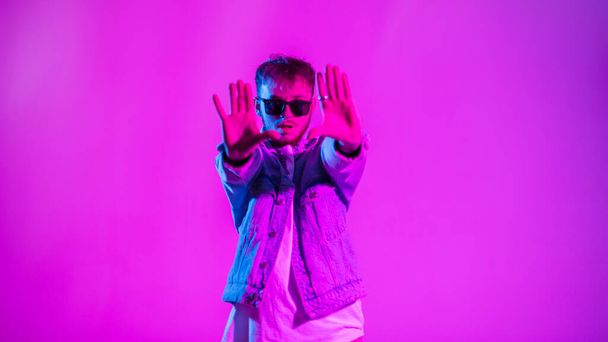 Knappe jonge hiphop danser man met stijlvolle zonnebril in fashion jeans jas dansen in kleurrijke studio met creatieve roze en blauw licht - Foto, afbeelding