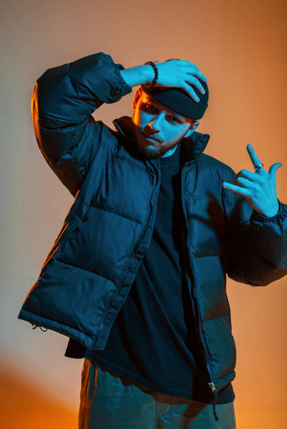 Fashionable handsome cool hipster man in stylish black clothes with down jacket adjusts his hat in creatine color studio with warm orange and cold neon light - Фото, зображення