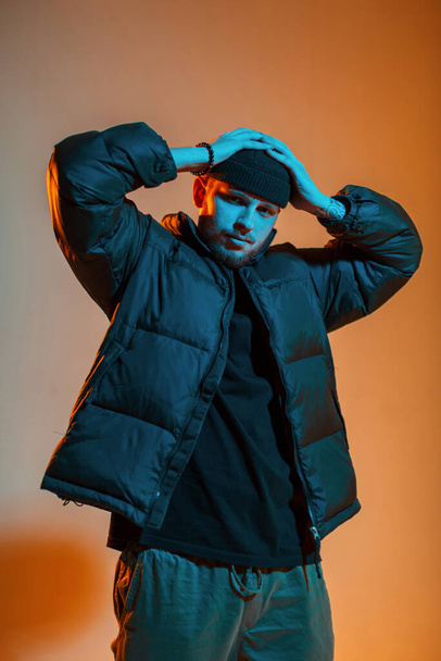 Cool stylish young handsome man in fashionable clothes with a winter down jacket puts on a hat and poses in a creative studio with orange and neon light - Фото, изображение