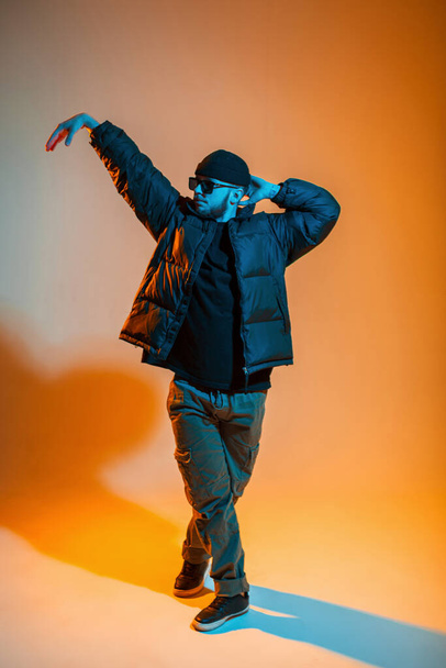 Stylish professional dancer man with sunglasses in fashionable clothes with a hat and a down jacket is dancing in a studio with creative orange and neon lights - Photo, image