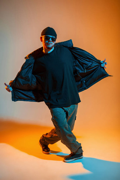 Módní profesionální pohledný muž se slunečními brýlemi a čepicí v módních černých šatech s bundou tančí v kreativním studiu s neonem a oranžovým světlem - Fotografie, Obrázek