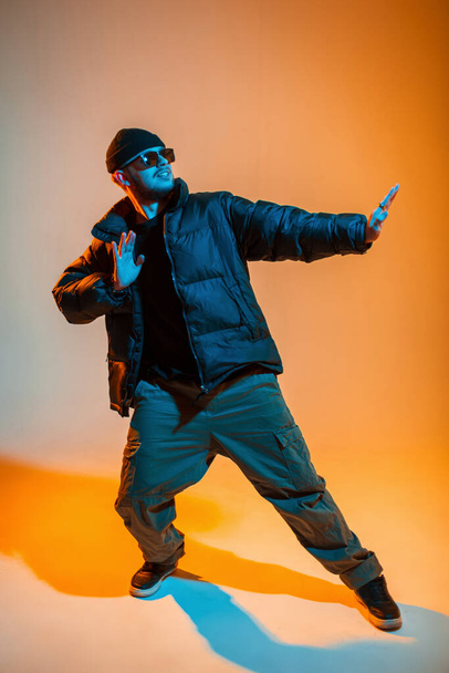 Elegante hombre guapo y moderno bailarín de hip hop profesional con gafas de sol y gorra en ropa negra de moda con chaqueta de plumón de invierno y zapatillas de baile en un estudio creativo con luz de neón naranja y fría - Foto, Imagen