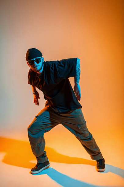 Beau jeune danseur professionnel avec des lunettes de soleil fraîches et une casquette dans un t-shirt noir de mode danse dans un studio lumineux créatif avec une couleur orange et néon - Photo, image
