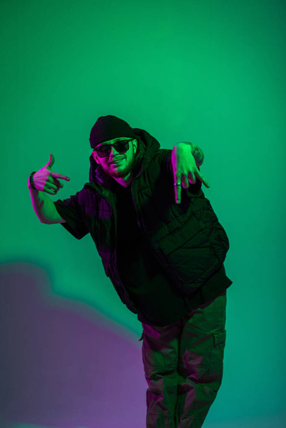 Elegante divertido hombre hipster de moda con gorra y gafas de sol en ropa de moda negro con chaleco posando en un estudio de color creativo con luz verde y púrpura - Foto, Imagen