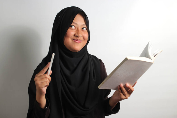 Excited young Asian girl in black hijab or headscarf holding a book and pen got an amazing idea isolated on green background - Photo, Image