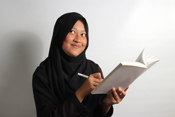 Jovem asiática excitada em hijab preto ou lenço de cabeça segurando um livro e caneta tem uma ideia incrível isolado no fundo verde - Foto, Imagem