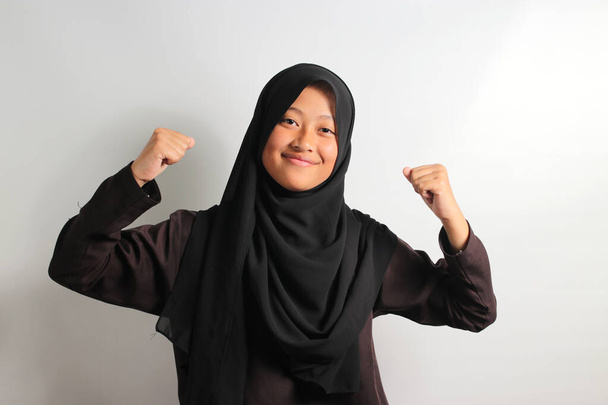 Podekscytowana młoda Azjatka ubrana w czarny hidżab lub chustę pokazującą silny gest podnosząc ramiona i mięśnie dumnie uśmiechając się do kamery odizolowanej na białym tle. - Zdjęcie, obraz