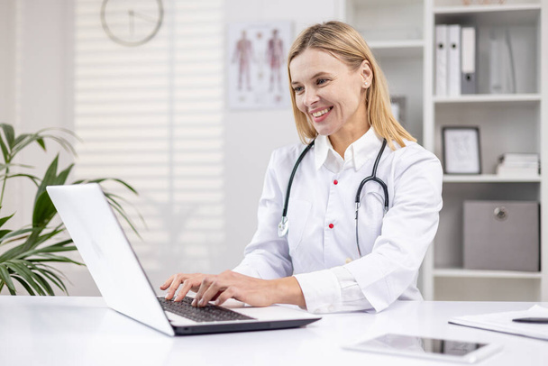 Mature female doctor working inside medical office, pediatrician using laptop while sitting inside clinic office, female worker in medical gown smiling. - Photo, Image