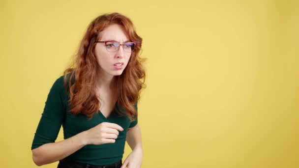 Рыжая женщина в очках с любопытством наблюдает в студии с желтым фоном - Кадры, видео