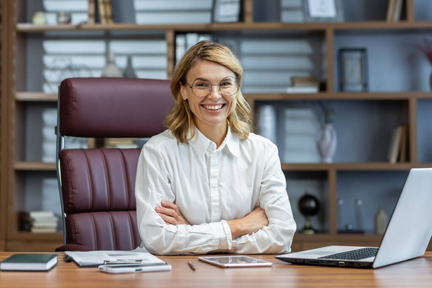 Portrait of an older beautiful blonde woman, director, boss, founder of the company in glasses and a white shirt. Sitting at the desk in the office, working on a laptop and smiling at the camera. - Foto, imagen