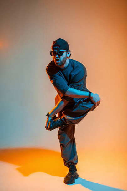 Havalı profesyonel hip hop dansçısı şık moda kıyafetleri içinde şapkalı ve güneş gözlüklü adam turuncu bir arka planda neon ışıklı duygularla dans ediyor. - Fotoğraf, Görsel