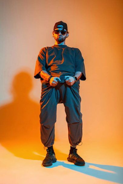 Cooler stylischer Hipster mit Sonnenbrille und Mütze in trendiger Mode, der sich an der Hose festhält und in einem kreativen Studio mit orangefarbenem und neonfarbenem Licht posiert - Foto, Bild