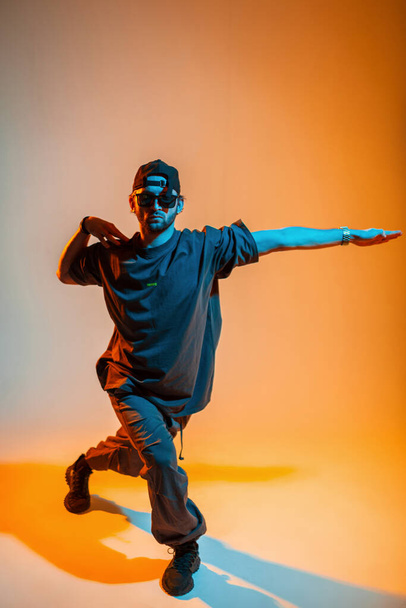 Modelo de cara profissional de moda legal com óculos de sol e um boné em uma camiseta, calças e tênis está dançando em um estúdio de concreto com luz laranja e néon - Foto, Imagem