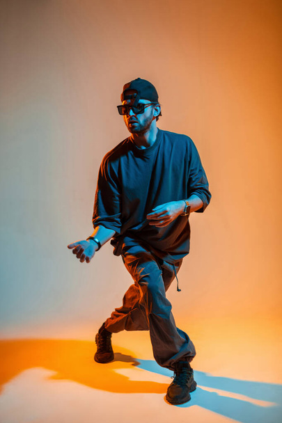 Stylowy moda hip hop tancerz człowiek w okularach i czapce w modne ubrania taniec styl moda w kreatywnym studio z pomarańczowym i niebieskim światłem - Zdjęcie, obraz