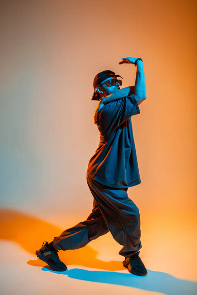 Stylischer gutaussehender Tänzer mit Mütze und Sonnenbrille in trendiger Kleidung tanzt in einem kreativen Studio mit orangefarbenem und neonfarbenem Licht - Foto, Bild