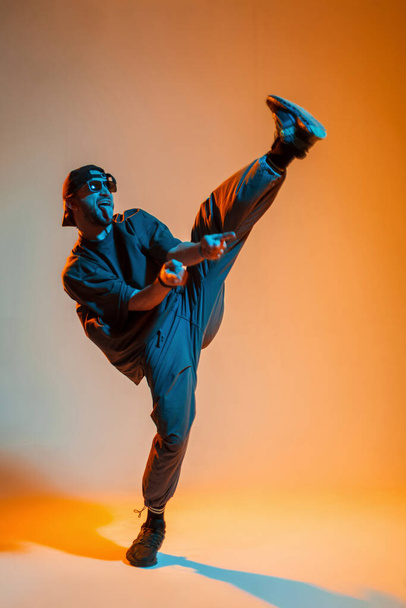Cool stylish fun handsome young dancer man with fashion sunglasses and a cap in fashionable outfit with a t-shirt is dancing in a creative color studio with orange warm and neon cold light - Foto, afbeelding
