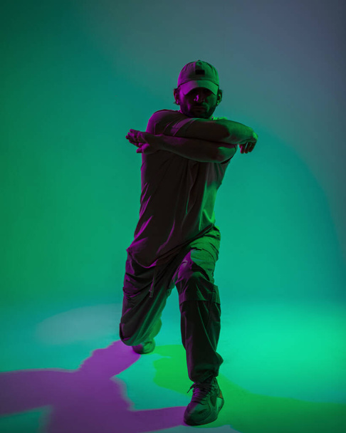 緑と紫のライトと創造的なスタジオで踊るファッション服のキャップとファッショナブルなハンサムなプロの男性ダンサー - 写真・画像