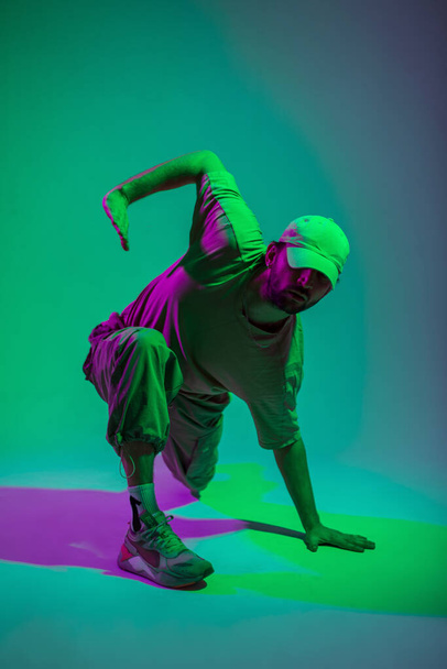 Cool hombre bailarín de hip hop profesional de moda con gorra en ropa de moda bailando en un colorido estudio con luces verdes y rosadas - Foto, imagen