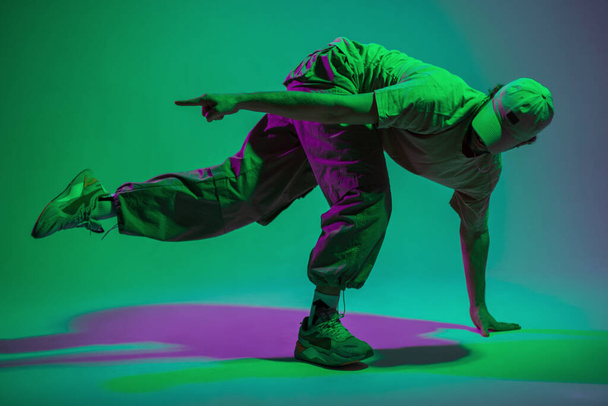 Moda hombre bailarín profesional con una gorra en ropa elegante con zapatillas de deporte bailando en el suelo en un colorido estudio creativo con luz púrpura y verde - Foto, imagen