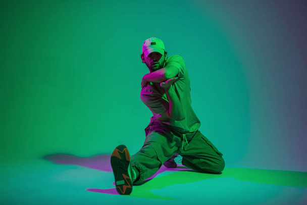 Módní pohledný profesionální tanečník muž s čepicí v módním oblečení sedí a tancuje v kreativním barevném studiu s azurovým a růžovým světlem - Fotografie, Obrázek