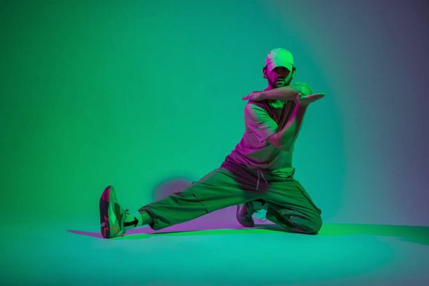 Moda przystojny profesjonalny tancerz człowiek z czapką i tenisówki w modne beżowe ubrania taniec i siedzi w kreatywnym studio kolor z cyjanu i światła magenta - Zdjęcie, obraz