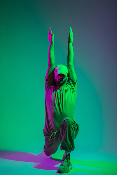 Cool danseur professionnel homme avec une casquette dans des vêtements élégants avec des baskets à la mode dansant dans un studio de couleur avec du cyan et de la lumière violette - Photo, image