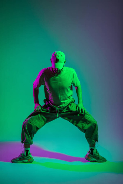 Cool creative fashion man dancer with cap in fashionable clothes with sneakers dancing in colored studio with cyan and magenta light - Photo, Image
