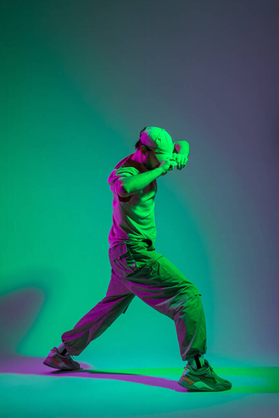 Hombre de bailarina profesional guapo de moda en ropa de moda bailando y ajustando su gorra en un estudio creativo con luz cian y magenta - Foto, Imagen