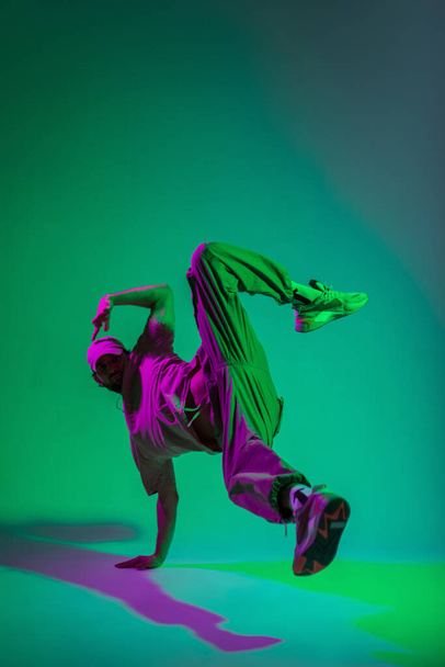 Stylowy modny profesjonalny tancerz z czapką w ubraniu modowym z tenisówkami tańczącymi w ruchu na rękach w kreatywnym kolorowym studio z różowym i fioletowym światłem - Zdjęcie, obraz