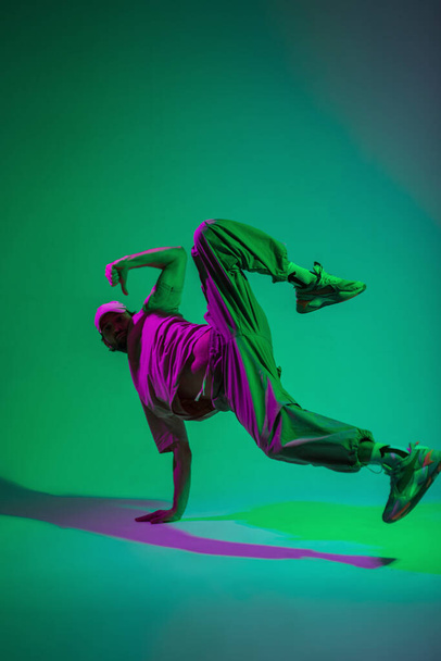 Stylish professional dancer guy hip hop b-boy in fashionable clothes with a cap and sneakers dances on hand in a creative studio with magenta and cyan color - Foto, imagen