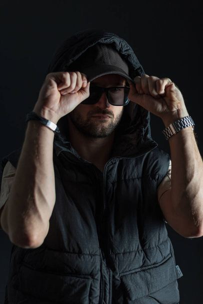 Fashionable handsome man with sunglasses in a fashion trendy hooded vest puts on a cap in the dark - Фото, зображення