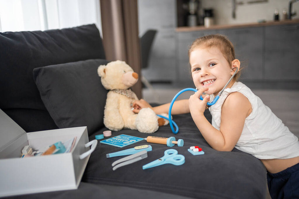 Pieni tyttö leikkii lääkäriä leluilla ja nallekarhu sohvalla olohuoneessa kotona. Laadukas kuva - Valokuva, kuva