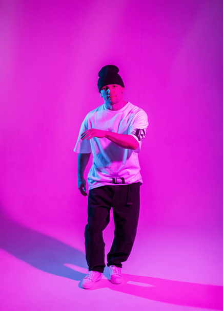 Jóképű hip-hop b-boy táncos divat ruhák egy kalap táncol egy színes stúdió rózsaszín és kék fény - Fotó, kép