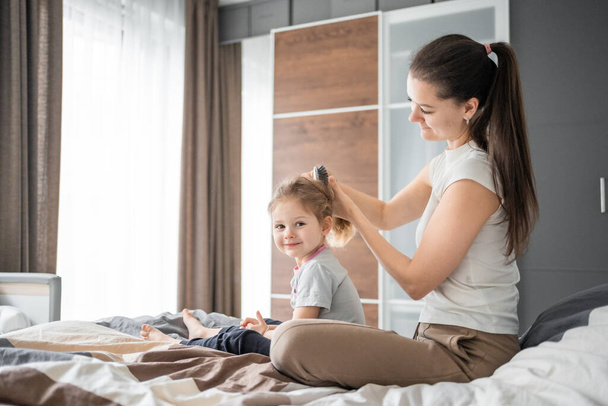 A jovem mãe penteia o cabelo de suas filhas enquanto se senta na cama pela manhã. Foto de alta qualidade - Foto, Imagem