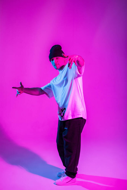 Cool stylish b-boy dancer guy in trendy clothes with a hat posing and dancing in a creative studio with pink and neon lights - Фото, изображение