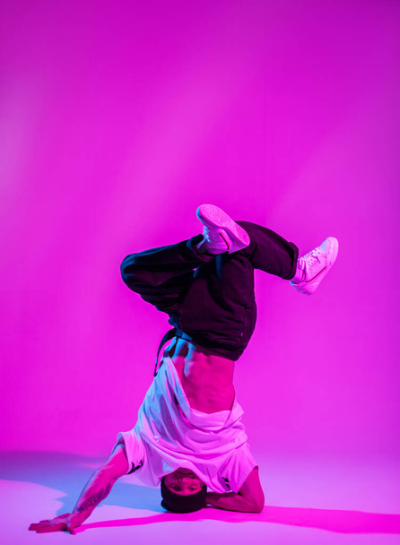 Stylowy profesjonalny tancerz b-boy w ubraniach modowych z tenisówkami tańczy i stoi na głowie w jasnym studio kreatywnym z różowymi i neonowymi światłami - Zdjęcie, obraz