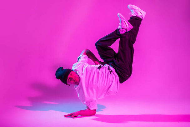 Professional breakdancer guy in fashion clothes with hat, t-shirt, pants and shoes dance and stands on hand in creative colorful studio with pink and neon lights - Photo, image