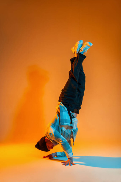 Stylish trendy professional breakdancer guy in trendy fashion clothes with a shirt is dancing on his hands in a creative studio with yellow and blue light - Fotó, kép