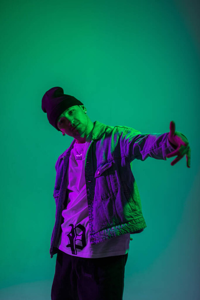 Cool fashionable artist hipster man with hat in denim stylish outfit poses in creative color studio with green and pink light - Zdjęcie, obraz