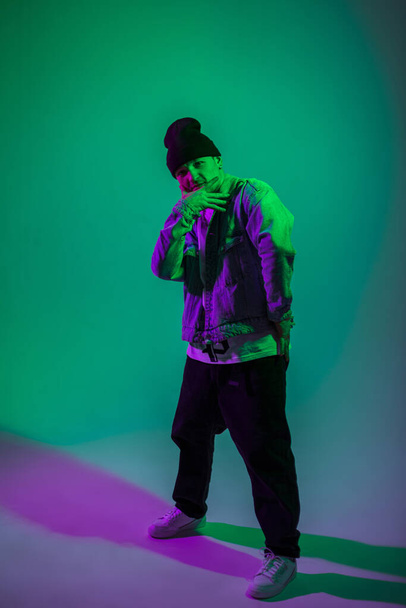Cooler stylischer trendiger B-Boy Tänzer in modischer Jeanskleidung posiert im Studio mit Cyan und Magenta Farbe - Foto, Bild