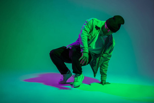 Módní pohledný mladý b-boy muž tanečník s čepicí v módní džínové oblečení tanec v kreativním studiu se zelenou a růžovou barvou - Fotografie, Obrázek