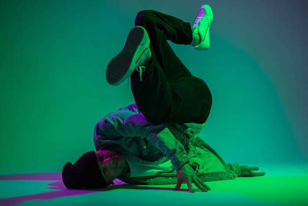 Cool fashion stylish hipster professional b-boy dancer in jeans clothes with denim jacket and sneakers dancing on the floor in a creative studio with purple and green light - Photo, Image