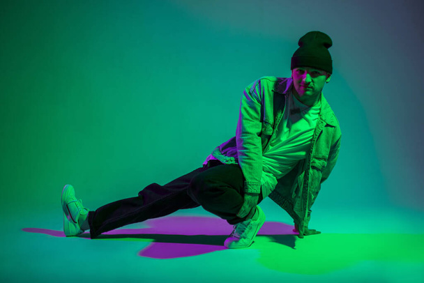 Bailarín profesional de moda b-boy hombre en traje de mezclilla elegante bailando y posa en el suelo en un estudio creativo con color verde y púrpura - Foto, Imagen