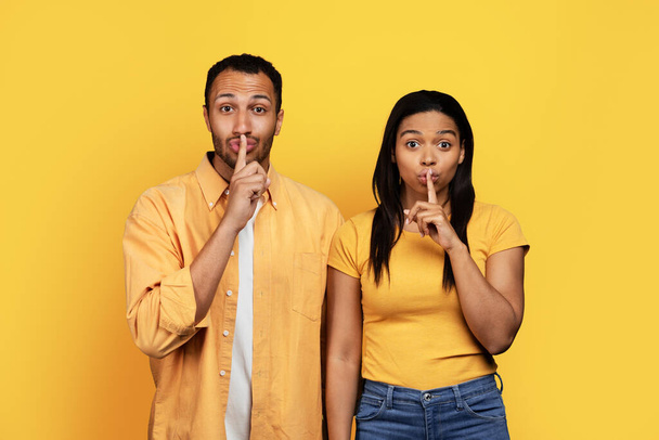 Millennial afroamericano chico y mujer poniendo el dedo en la boca, haciendo shhh signo aislado en el fondo del estudio amarillo. Secreto, misterio, sorpresa, emociones, anuncio y oferta - Foto, imagen