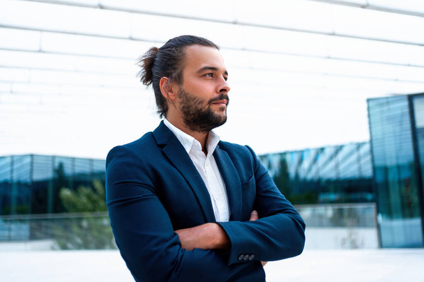 Successful businessman in suit with beard standing in front of office glass building arm crossed confidently looking away. Hispanic male business person side view portrait. Free space. Hipster man - Valokuva, kuva