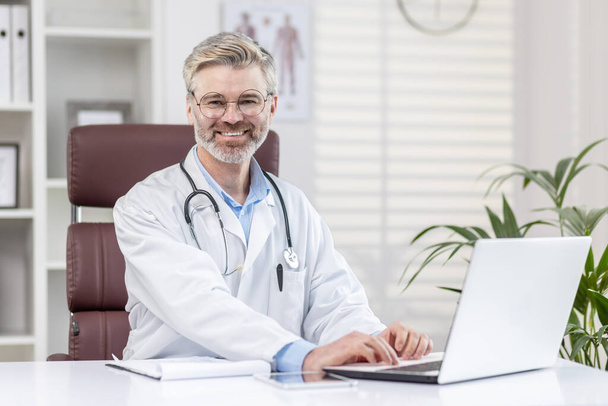 Portrait of a mature adult gray-haired doctor inside a medical office, the doctor is smiling and looking at the camera, the man is sitting at a table with a laptop and in a white medical coat. - Foto, Imagem