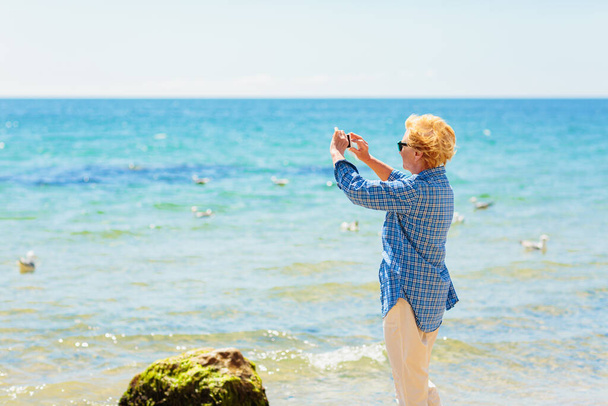 Elderly woman standing on the beach by the sea and taking photo of the sea and seagulls on mobile phone. Active seniors concept - Фото, зображення