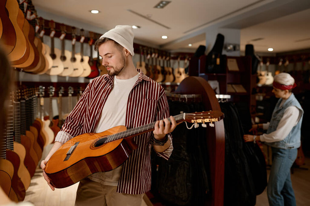 Satisfied man musician glad to choose new acoustic guitar for performance. Young salesman presenting improved model of classical string instrument. At music shop - Photo, Image